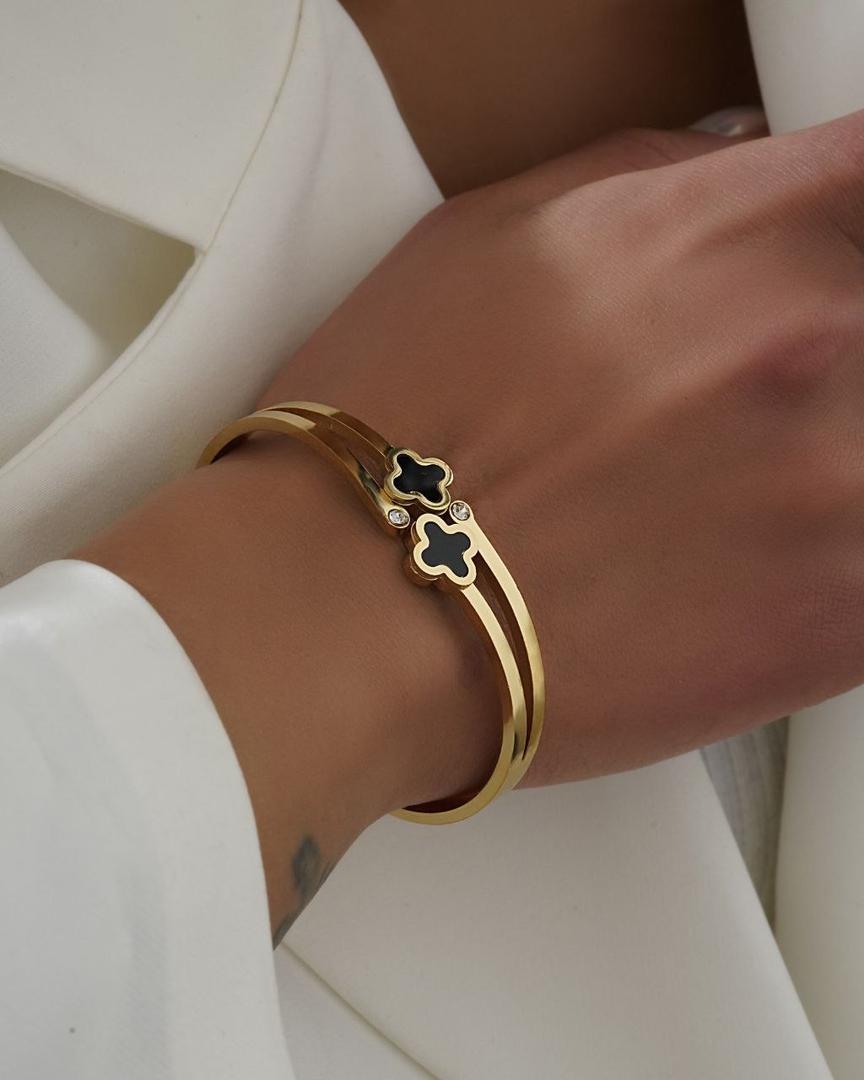 gold-bracelet-black-clover.jpg دستبند طلایی با طراحی دوخطی و آویز شبدر مشکی، تزئینشده با نگینهای درخشان، مناسب برای استایلهای لوکس و روزمره.”
